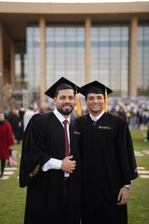Two males are standing and smiling. They are were wearing their graduation caps and gowns