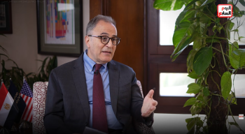 A man is sitting and talking. He is wearing a suit and glasses
