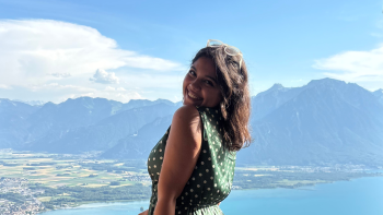 A girl standing in a balcony and behind her is a view of mountains, the sea, and greenery