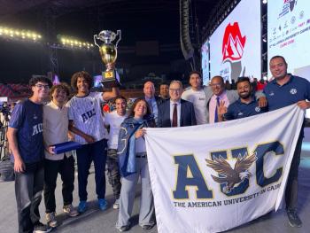 A group of AUC athletes stand with President Ahmad Dallal and Dean of Students Yorgen Marcel smiling, as one of them holds up the cup