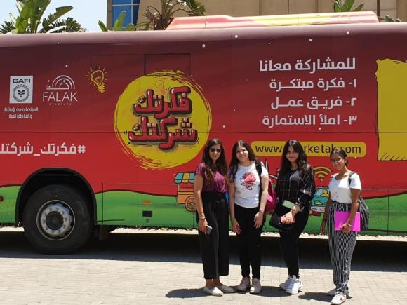 A picture of four girls in their twenties standing in front of a bus covered with texts and logos, and a main slogan that says "Your idea is Your company".