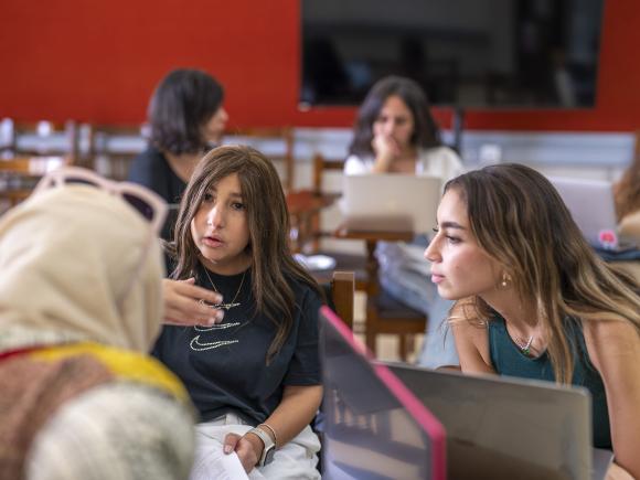 Two girls sitting in class with their laptops on the desk talking to their teacher