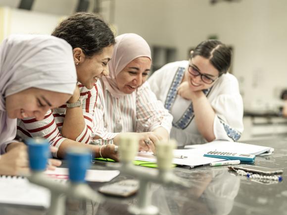 Three girls in a lab with their professor smiling and writing in their notebooks