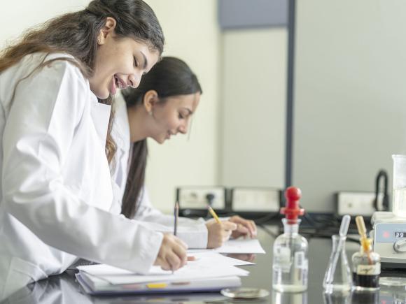 Two girls wearing white lab coat writing in their notebooks with test tubes on the table
