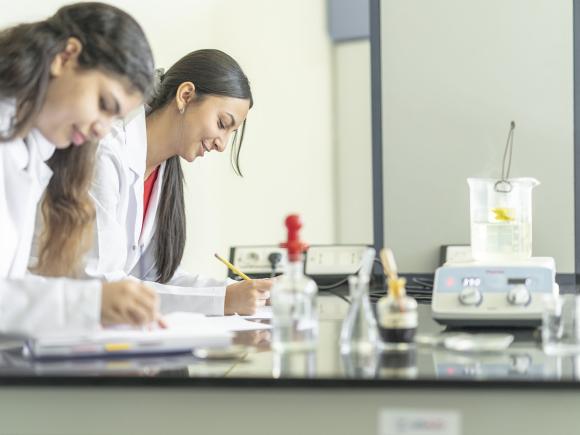 Two girls wearing a white lab coat writing in their notebooks in a lab