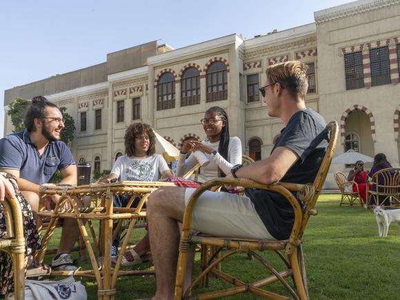 A group of girls and boys sitting outdoors around a table in a garden with an Islamic building behind them