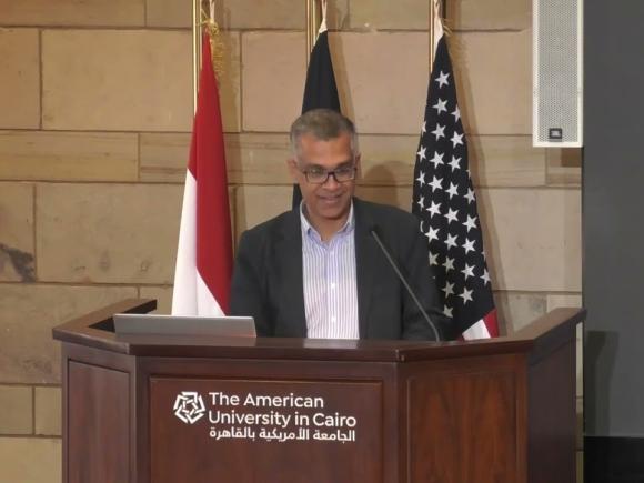 Man wearing a suit talking on a podium with 3 flags behind him