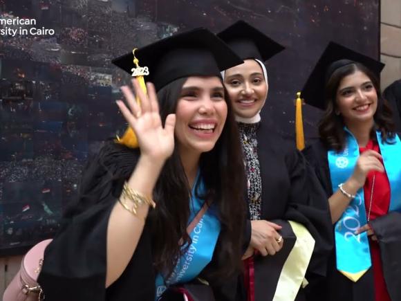 Three females are sitting and a male is standing and wearing their caps and gowns