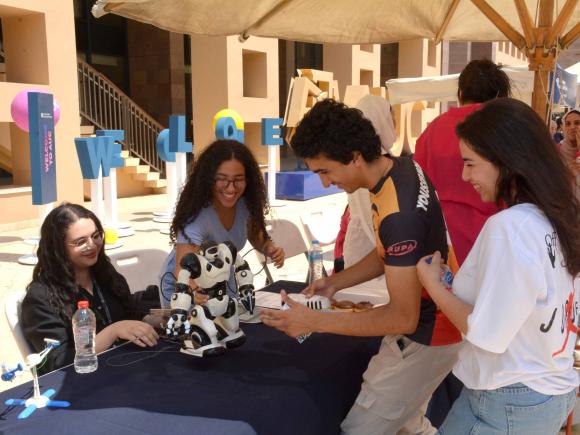 A group of girls and boys standing by a booth looking at a robot on the table