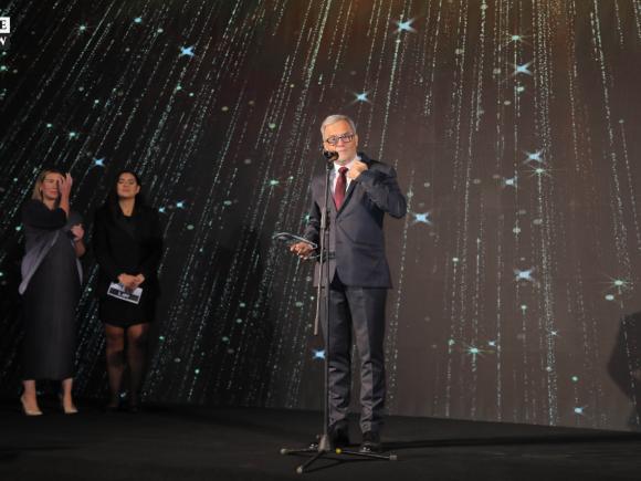 A man with a navy suit and a red tie stands in front of a conference background at a microphone, holding a glass award in his hand