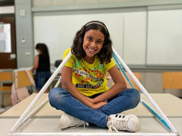 A young student sitting on a desk with a pyramid wrapped around her made out of resourceful materials