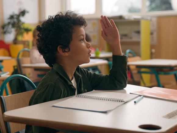 a young boy sitting on a desk in a classroom and raising his hand up