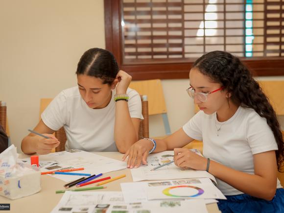 two young girls wearing white shirts sitting at a round table with colored pencils and papers