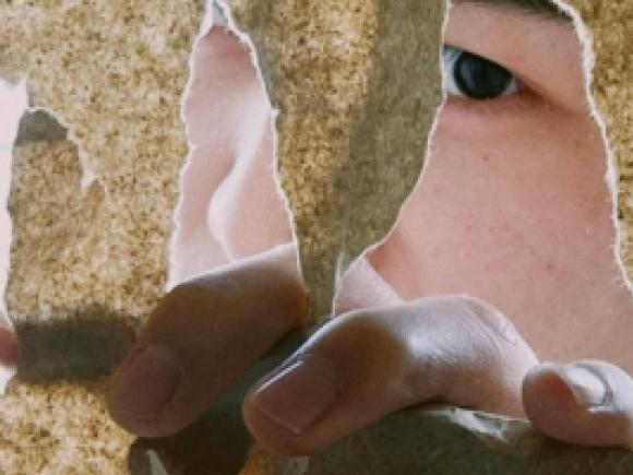 A close-up of a hand tearing through textured paper, revealing part of a person&rsquo;s face behind it.