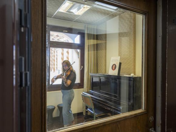 Student practicing violin in a music practice room with a piano and natural light from the window.