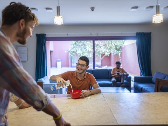 Students socialize in a residence hall common area while one prepares cereal in the shared kitchen.