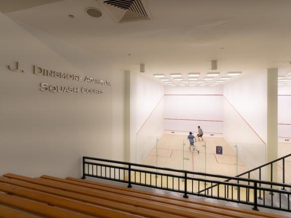 Two players compete in a squash match inside the J. Dinsmore Adams Jr. Squash Courts facility at AUC, viewed from spectator seating.