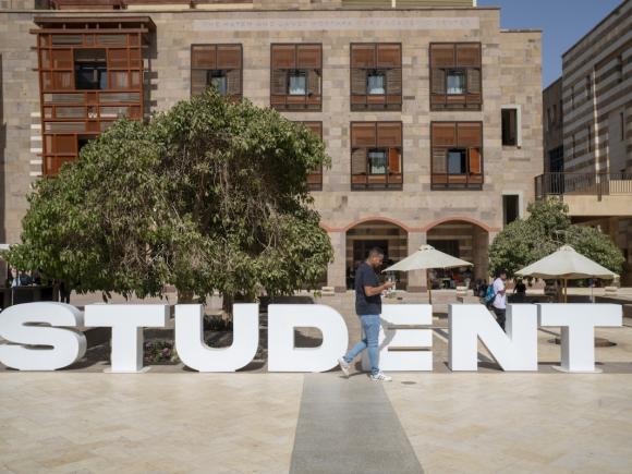 Large &ldquo;STUDENT&rdquo; sign in the courtyard of the Hatem and Janet Mostafa Core Academic Center with students walking nearby.