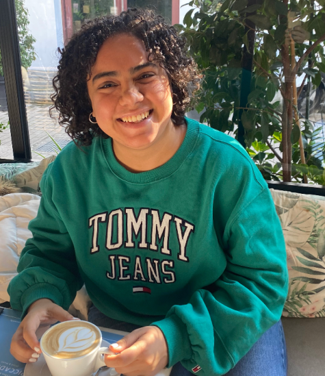 A girl is smiling and sitting indoor, holding a cup of coffee. The text on her sweatshirt reads "Tommy Jeans"