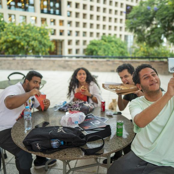 Group of girls and boys sitting outdoors taking a selfie while eating