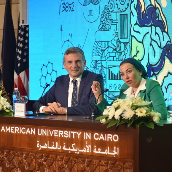 A man and a woman are sitting on a panel and the woman is talking in a microphone