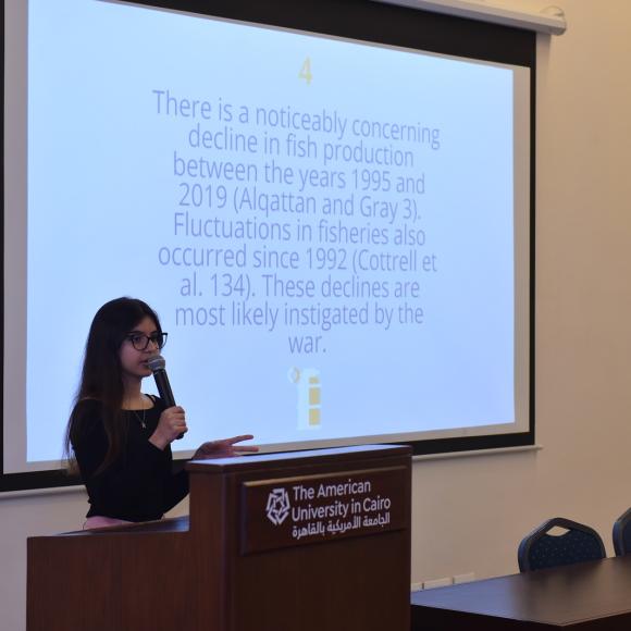 A female is talking in a microphone in front of a screen displaying text