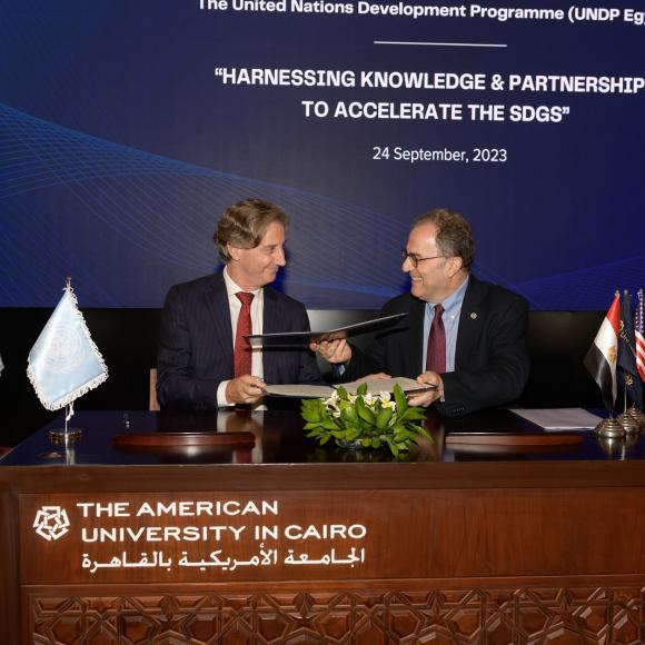 Two men handing leather files to each other, text reads "The American University in Cairo الجامعة الأمريكية بالقاهرة"