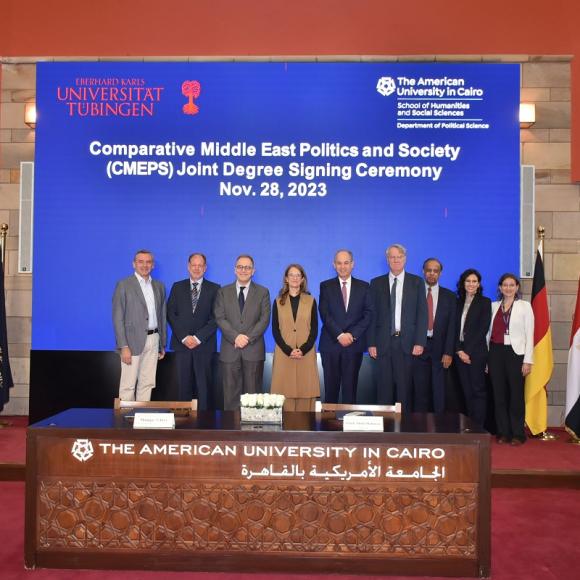 People standing next to each other and in between flags on a podium, text reads "Comparative and Middle East Politics and Society (CMEPS) Joint Degree Signing Ceremony"