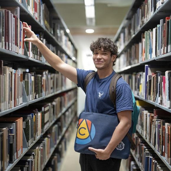 A male is smiling and standing in front of big book shelves reaching for a book and holding a laptop sleeve that reads the letters AUC