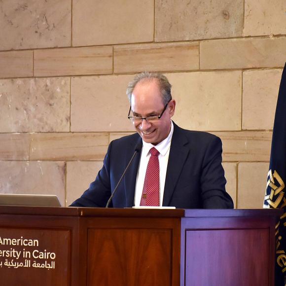 a man with gray hair wearing a suit and a red tie giving a speech
