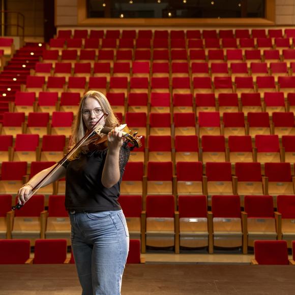 A female wearing glasses playing the violin in front of stage seats