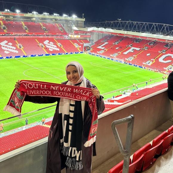 A veiled girl is smiling and standing in a stadium. She is holding a scarf with the text "Liverpool You'll Never Walk Alone, Liverpool Football Club"