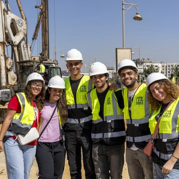 A group of males and females wearing vests and protective helmets and standing in a construction site
