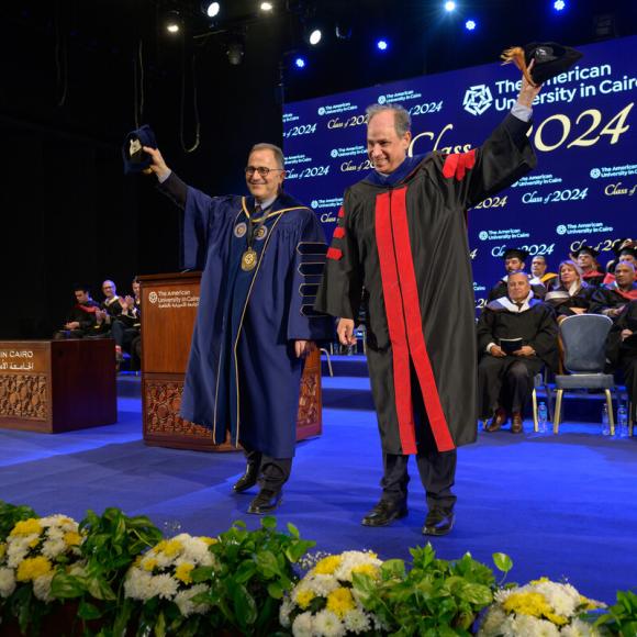 Two men are on stage raising their caps, the people behind them are seated and wearing caps and gowns.Text: The American University in Cairo.Class of 2024