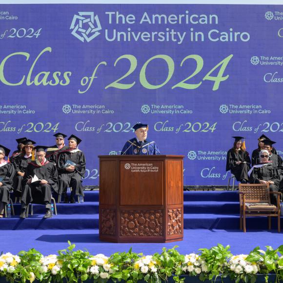 A man is standing at a podium, the people next to him are weraing caps and gowns: Text: The American University in Cairo.Class of 2024