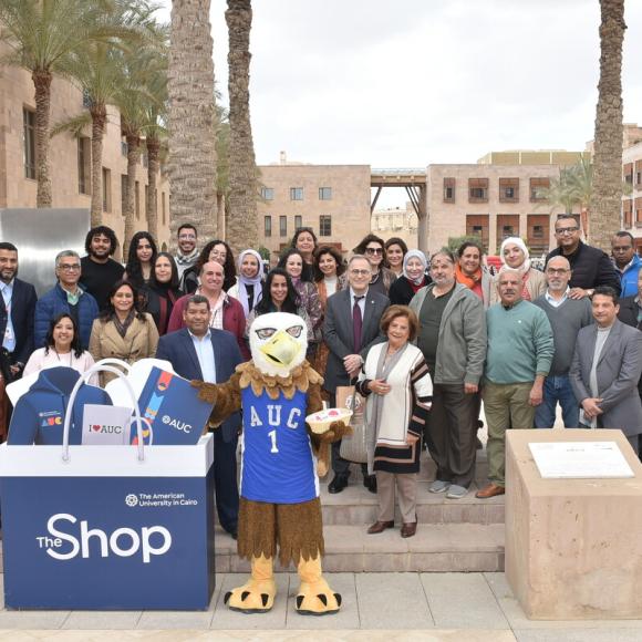A group of people are standing behind an eagle mascot and a big bag with the text: The Shop