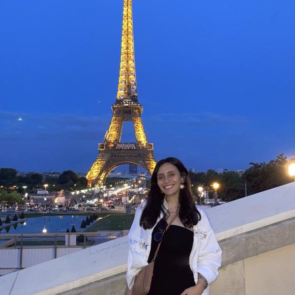 A female is standing and smiling. The Eiffel Tower is behind her.