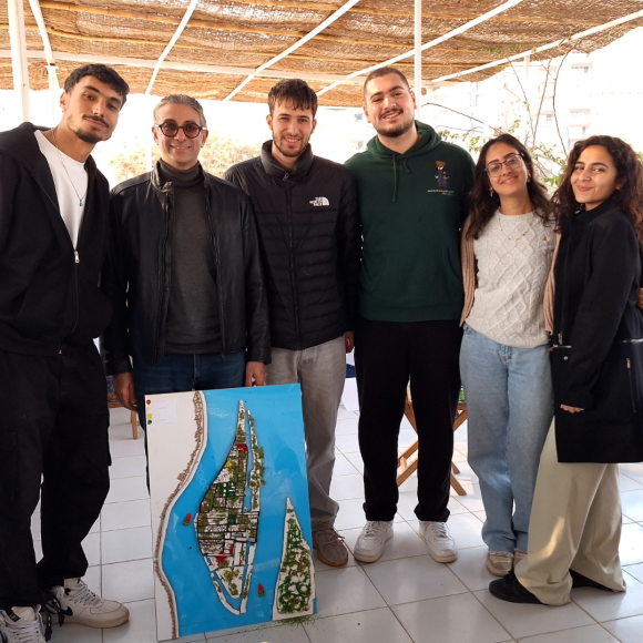 A group of students and faculty smiling and standing together on Qursaya Island next to an urban design poster the students created
