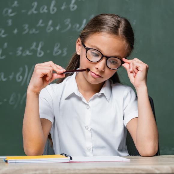 a young girl wearing a white tshirt and black eye glasses sitting on a desk holding a pen in her mouth