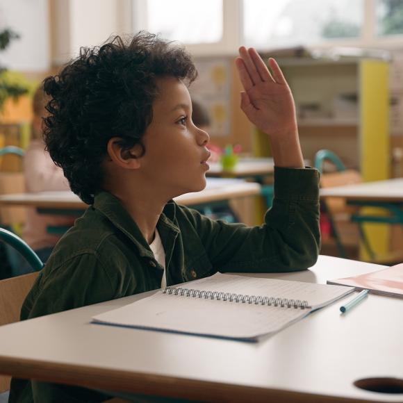 a young boy sitting on a desk in a classroom and raising his hand up
