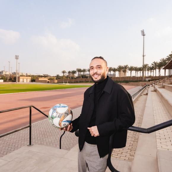 A man is standing in a field, he is smiliong and holding a football in his hand