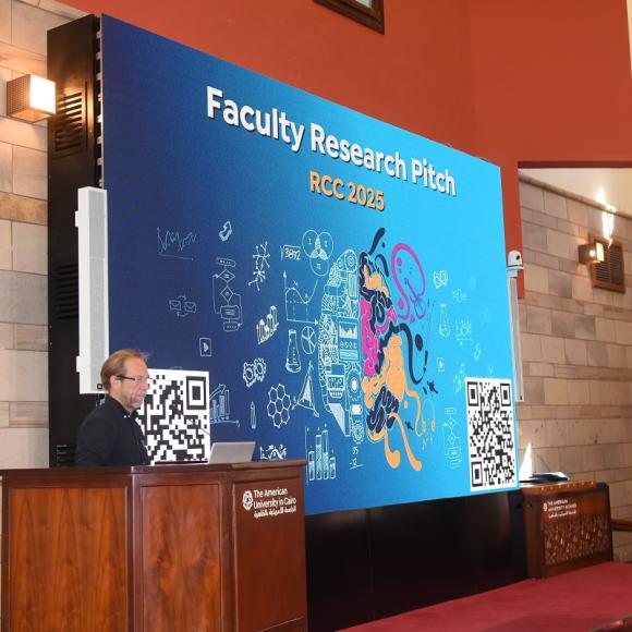 A man is standing at a a podium. Text: Faculty Research Pitch