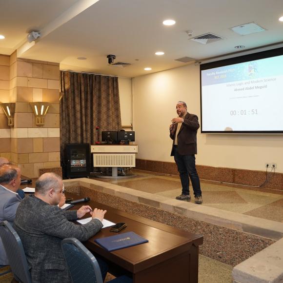 A man is talking into a microphone in front of a panel. Text: Faculty Research Pitch. Islamic Logic and Modern Science. Ahmed Abdel Meguid