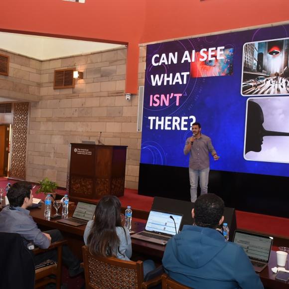 A man is talking in front of a panel. Text: Can AI See What Isn't There?