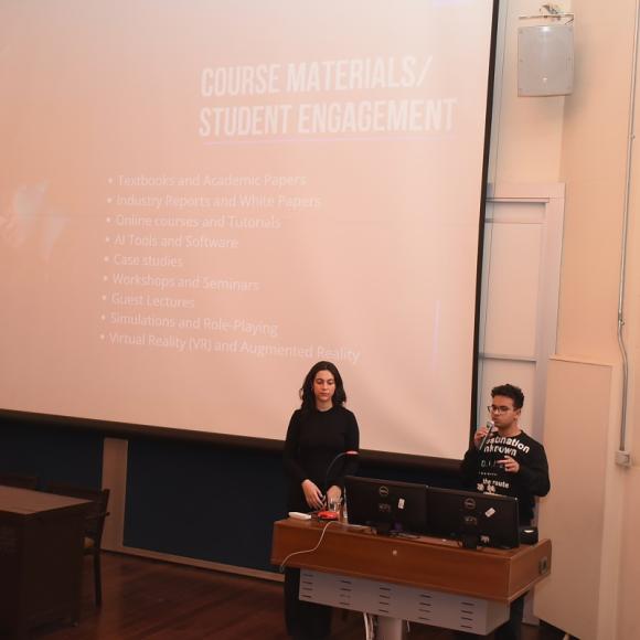 A male and a female are standing in front of a big screen. The male is talking into a microphone. Text: Course material. Student Engagement