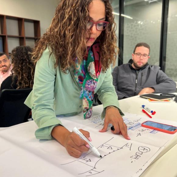 a woman with curly brown hair wearing glasses and a mint blouse holding a marker and writing on a paper