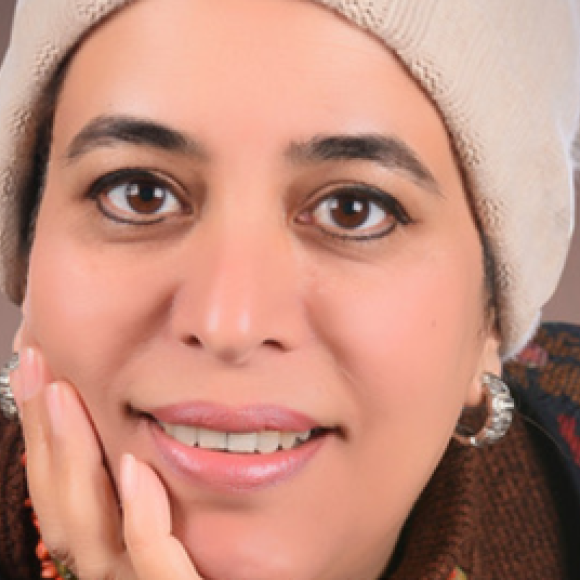 a headshot of a woman wearing a beige headscarf with her hand on her cheek