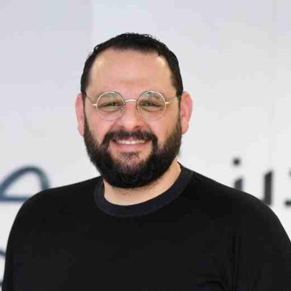a headshot of a man with black hair and black beard wearing glasses