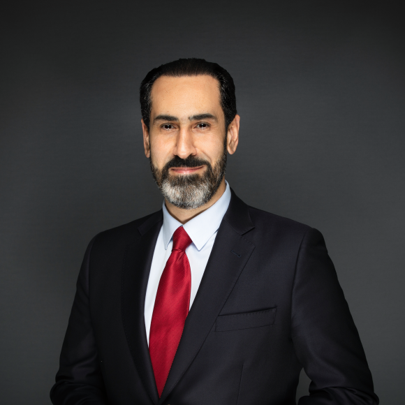 a headshot of a man wearing a black suit and a red tie with black hair and a grey beard