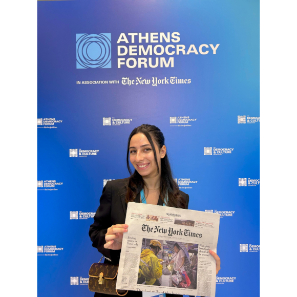 Laila Khalifa standing in front of a Athens Democracy Forum banner holding a copy of the newspaper the New York Times